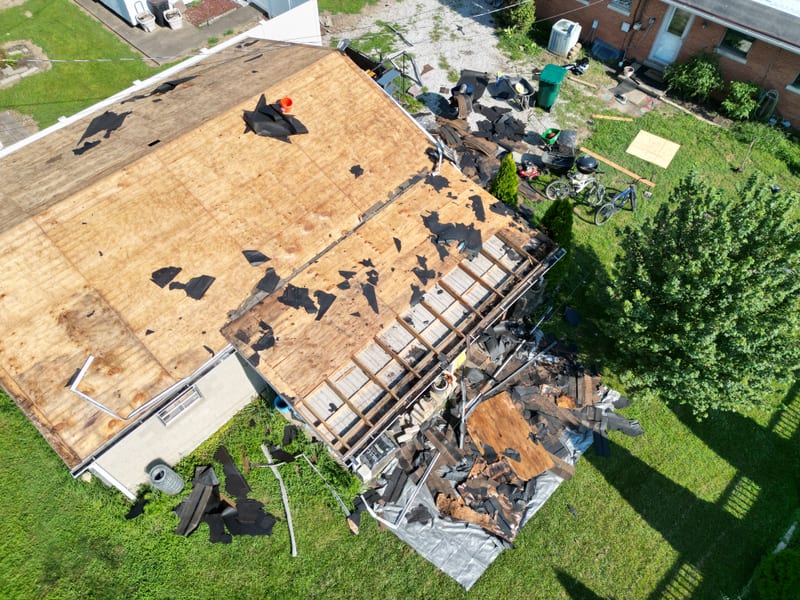 Top-down drone view of one roof stripped to bare decking with tear-off debris on tarps in the yard