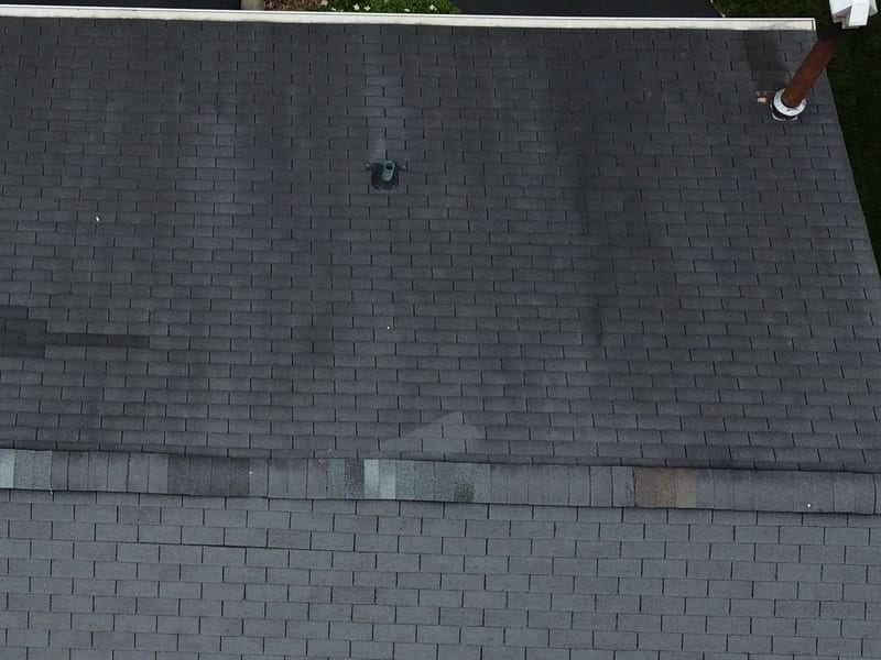 Aerial drone view of an asphalt shingle roof: the upper north-facing plane is covered in dark algae streaks running down the slope while the plane on the other side of the ridge is still clean