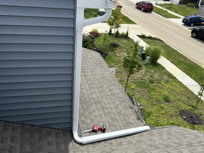 Rooftop view of a new gutter corner and downspout at the eave, overlooking the neighborhood street