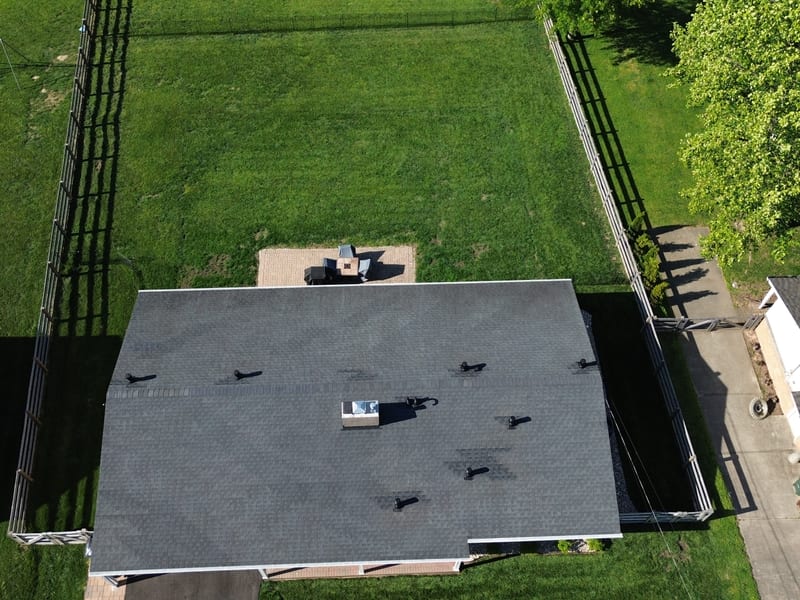 Overhead drone view of a gray shingle roof with seven new low-profile vents and turbines installed, large fenced backyard below