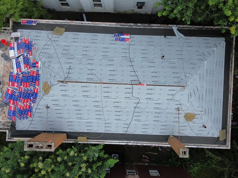 Top-down view of underlayment day: synthetic underlayment field with staged shingle bundles and safety lines on the duplex roof