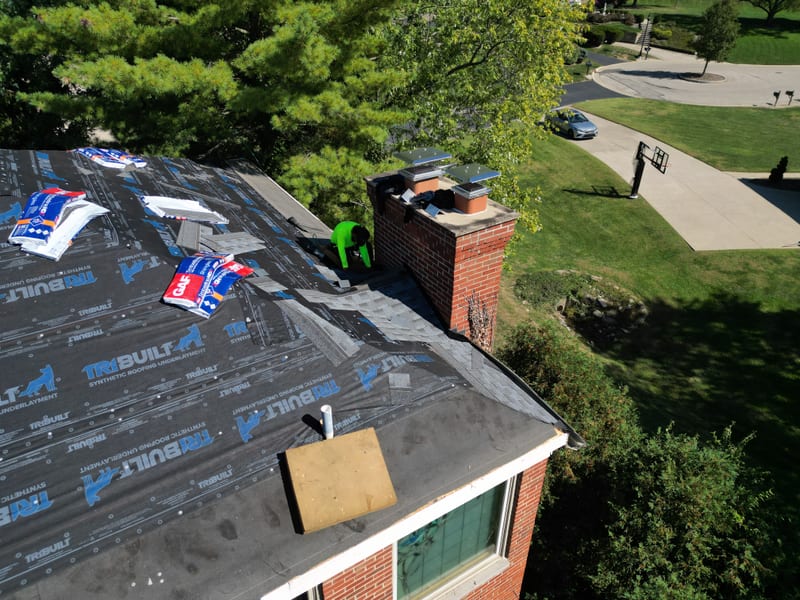Underlayment-covered plane with a roofer detailing the brick chimney and shingle bundles nearby