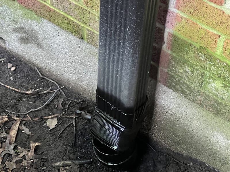 Close-up of a new black downspout base connector fitted where the downspout meets the foundation