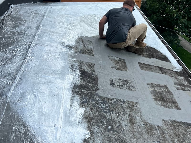 Fresh silver coating going down over the primed low-slope membrane, seen from behind the working roofer