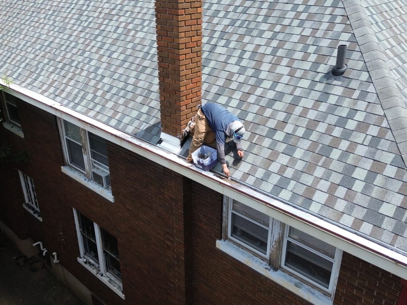 A roofer nailing off beside the brick chimney as the new shingle courses reach it