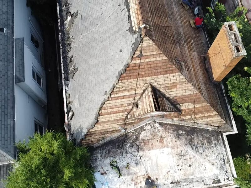 Top-down view of exposed plank roof decking and the dormer opening beside remaining old shingles and a brick chimney