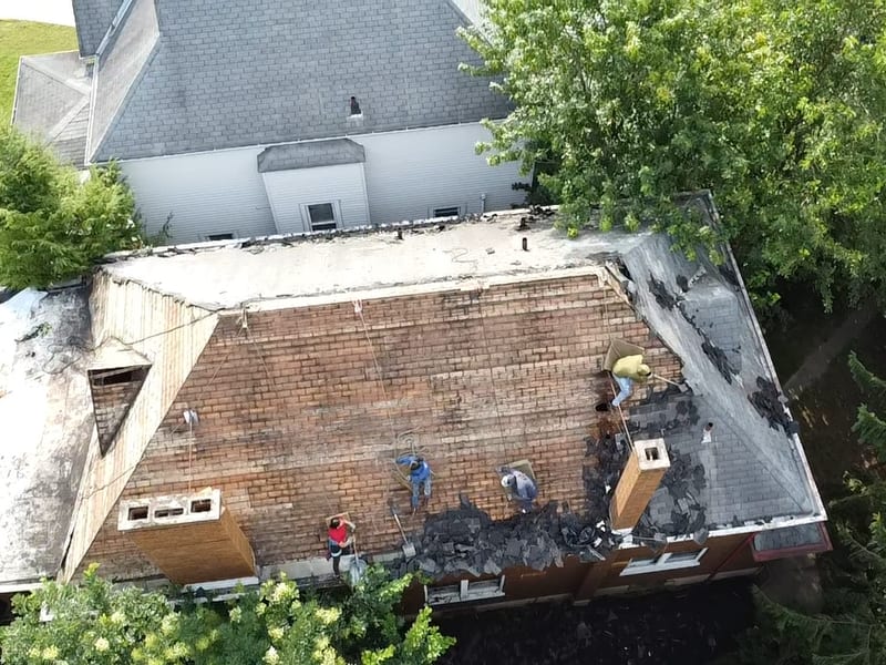 Wide drone view of the crew stripping the hip roof to plank decking, old shingles piling at the eaves