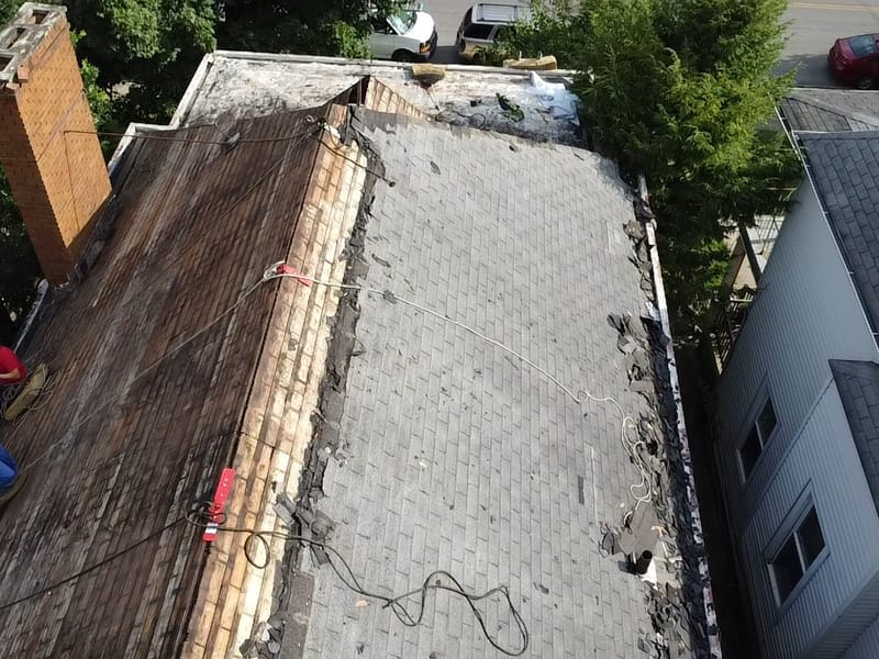Half-stripped roof from above: original plank decking exposed beside the brick chimney with crew working