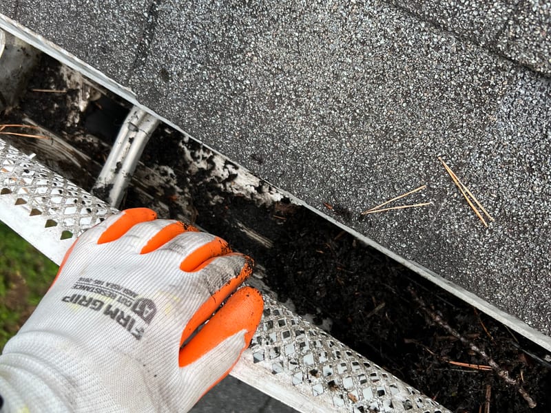 Gloved hand clearing packed leaf debris out of a gutter run by hand, mesh guard lifted for access