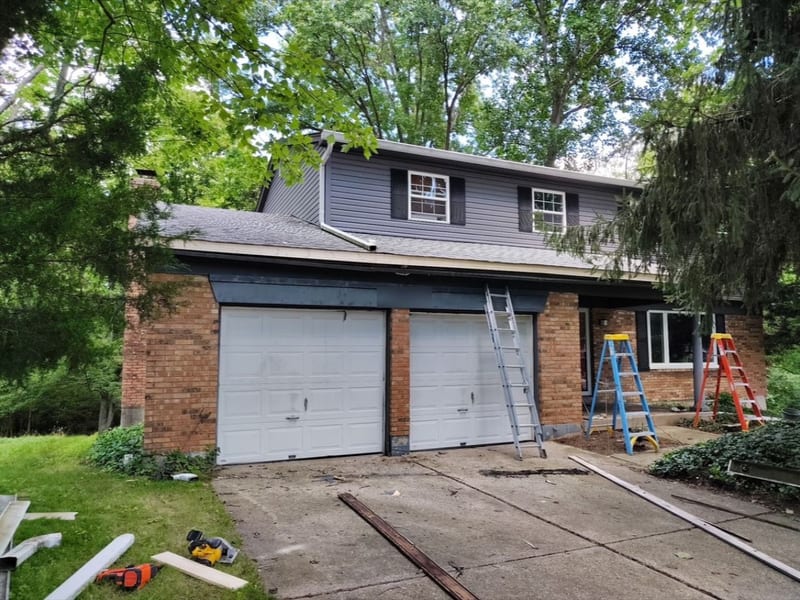 Two-story brick and siding home mid-installation with ladders set and the old gutter removed above the garage