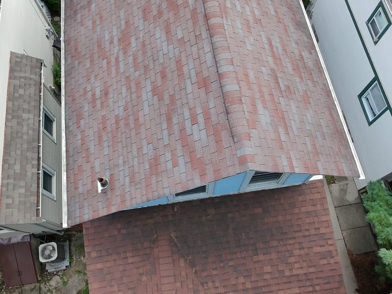 Aerial view of a shingle roof between two neighboring houses, showing the ridge, chimney vent, and dormer window