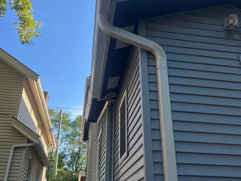 Ground-level view of a new white gutter and downspout running down the corner of a blue-sided house