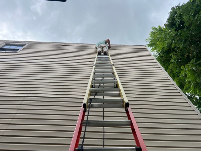 Extension ladder reaching a third-story gable while a technician re-secures a piece of vinyl siding at the top