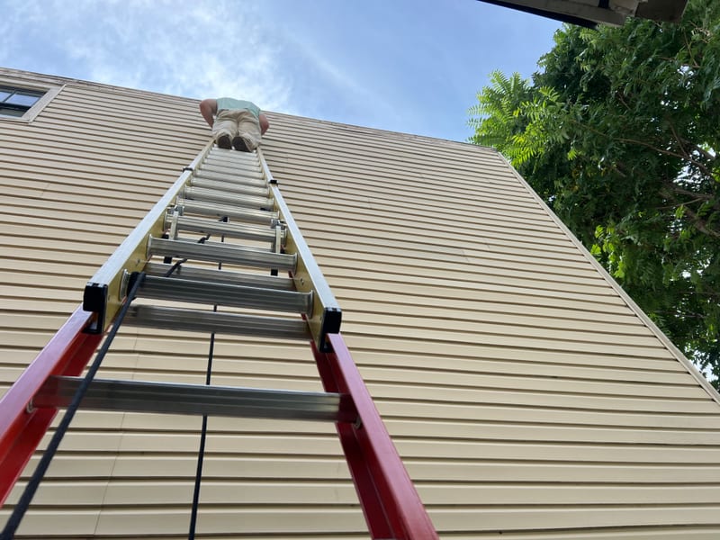 View up the extension ladder to the tan vinyl siding gable during the same-day repair