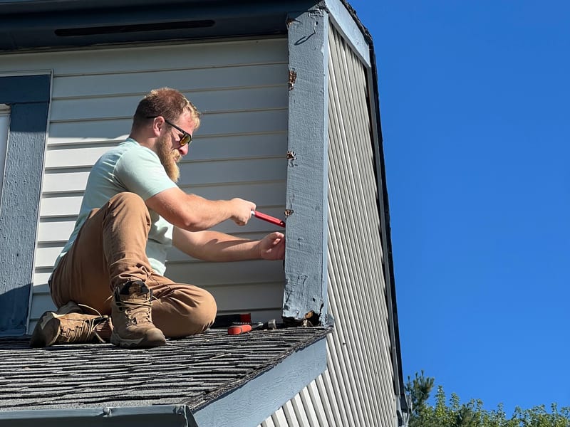 Joe kneeling on a roof edge removing a damaged corner trim board with a pry tool