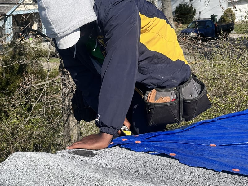 Crew member fastening the blue emergency tarp along the ridge with orange cap nails