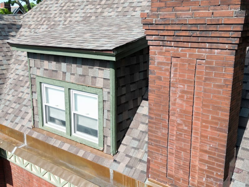 Close detail of new shingles running into a green-trimmed, shingle-sided dormer beside the chimney