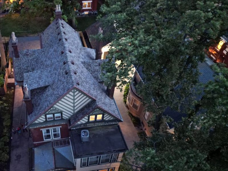 Dusk aerial of the whole house, the new roof reading pale against deep summer tree canopy