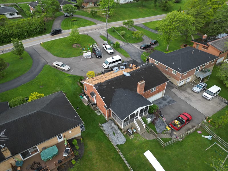 High aerial over the backyard toward the cul-de-sac with the crew working on exposed decking mid tear-off