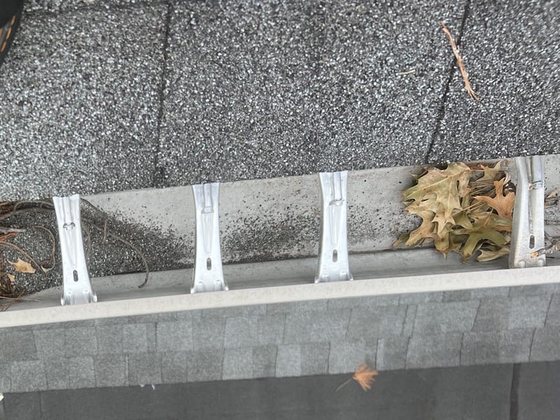 Looking down at a gutter trough from the roof edge, with shingle grit, twigs, and a clump of dried oak leaves collecting between the hanger brackets