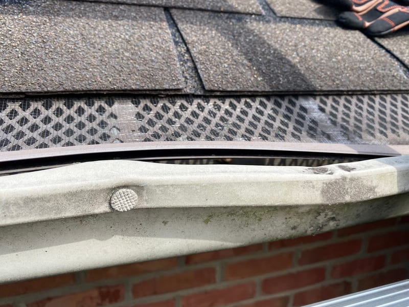 Close-up of an aged gutter with a bent front lip, a spike fastener backing out, and a worn mesh gutter screen no longer sealing against the shingles
