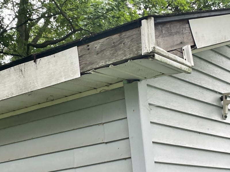 Corner of a roofline with the gutter missing, exposing rotted fascia board and a sagging soffit panel