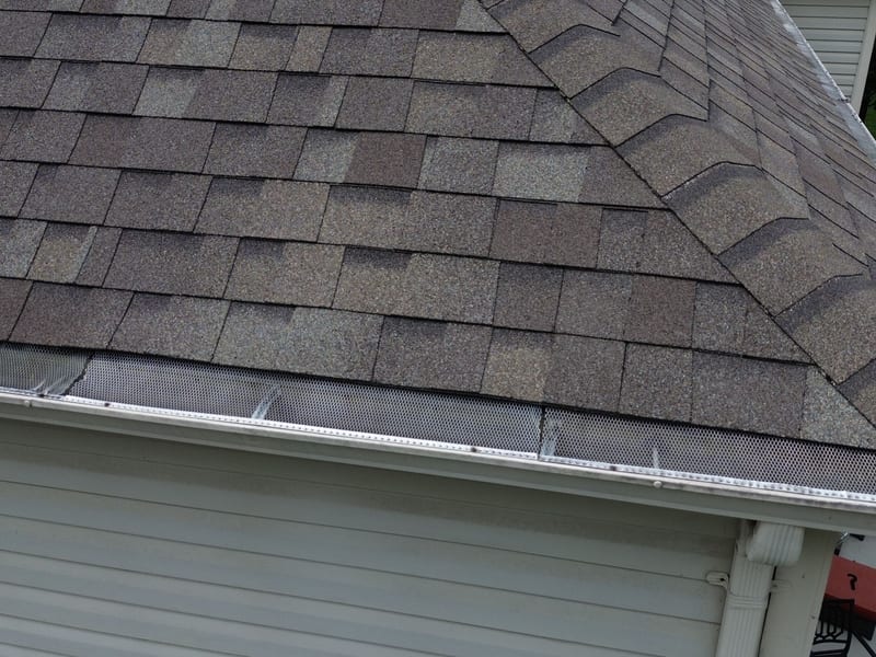 Drone close-up of expanded-metal gutter screens seated flat along a white seamless gutter run beneath architectural shingles at a hip corner