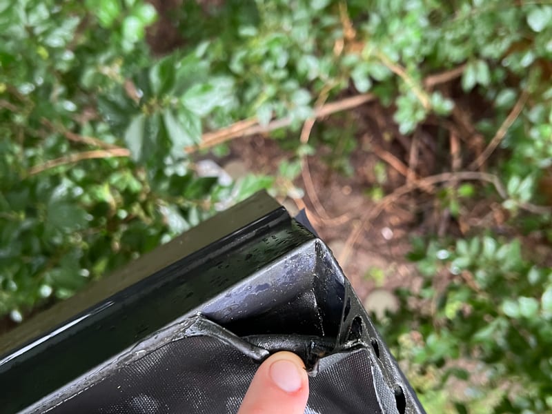 Fingertip pointing to torn, bunched EPDM membrane at a corner drain fitting on a flat roof edge, where the sheet should lap flat and sealed