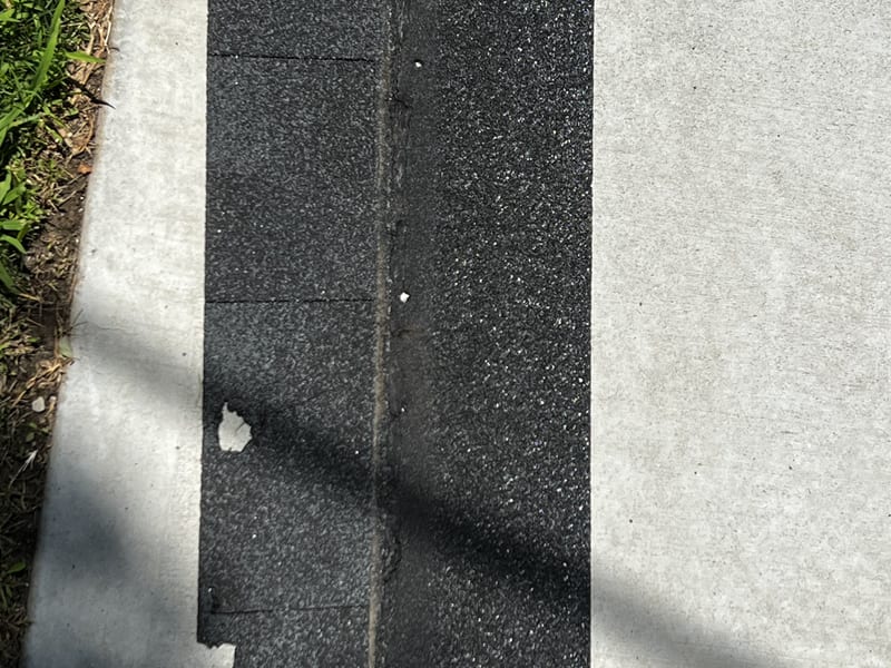 Full strip of aged three-tab asphalt shingle laid on a concrete walkway after removal from the roof, showing heavy granule loss, a worn seam line, and torn spots along one edge
