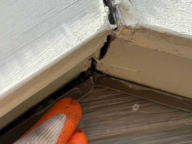 Close-up before photo at a gable peak: a gloved hand points to an open gap where the painted fascia trim and J-channel meet above vinyl siding