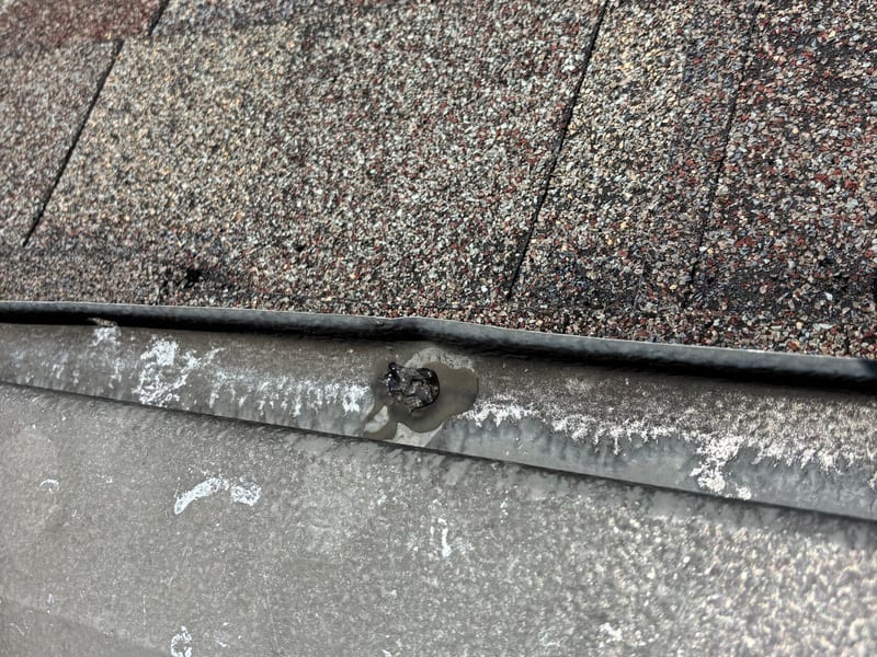 Detail of a metal flashing seam on an asphalt shingle roof, with an exposed fastener head capped under a dab of black sealant
