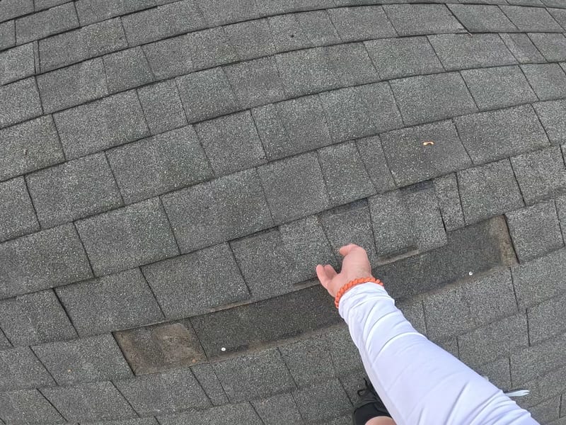 Close-up of a roofer’s hand pointing to missing shingle tabs and exposed dark underlayment on a wind-damaged asphalt roof