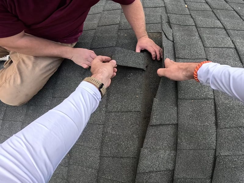 Two roofing technicians examine a delaminated shingle tab lifted from the roof, showing the torn edge left by wind damage