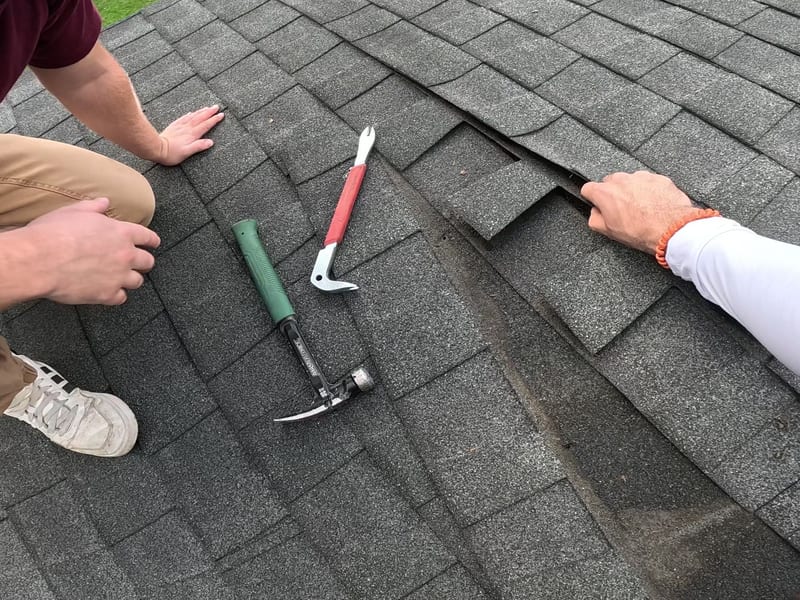 Hammer and nail-puller pry bar laid on the shingles as a technician lifts the edge of a wind-damaged shingle to begin replacement
