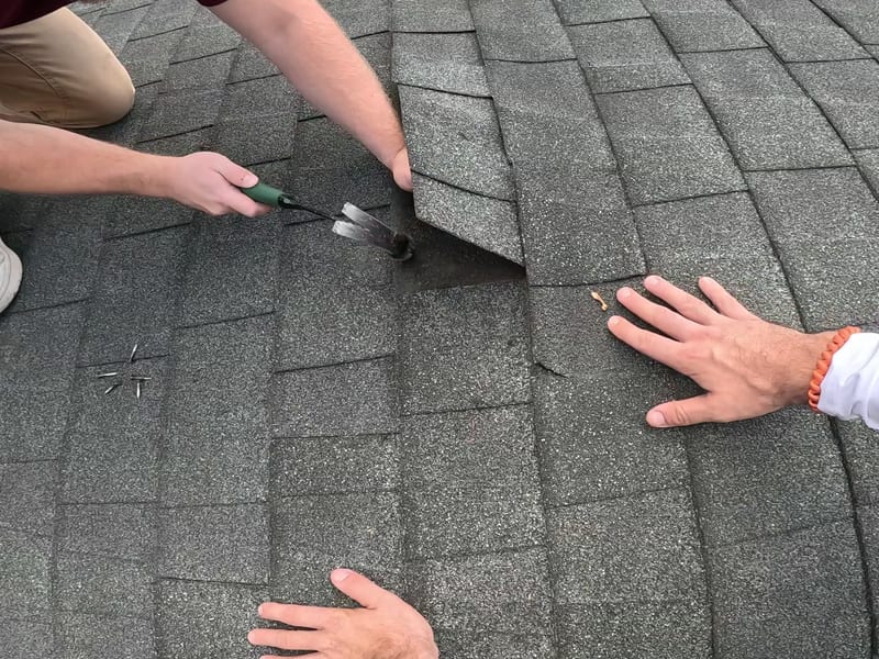 Technician uses a nail puller to extract fasteners from beneath a lifted shingle, with pulled roofing nails lying on the shingles nearby