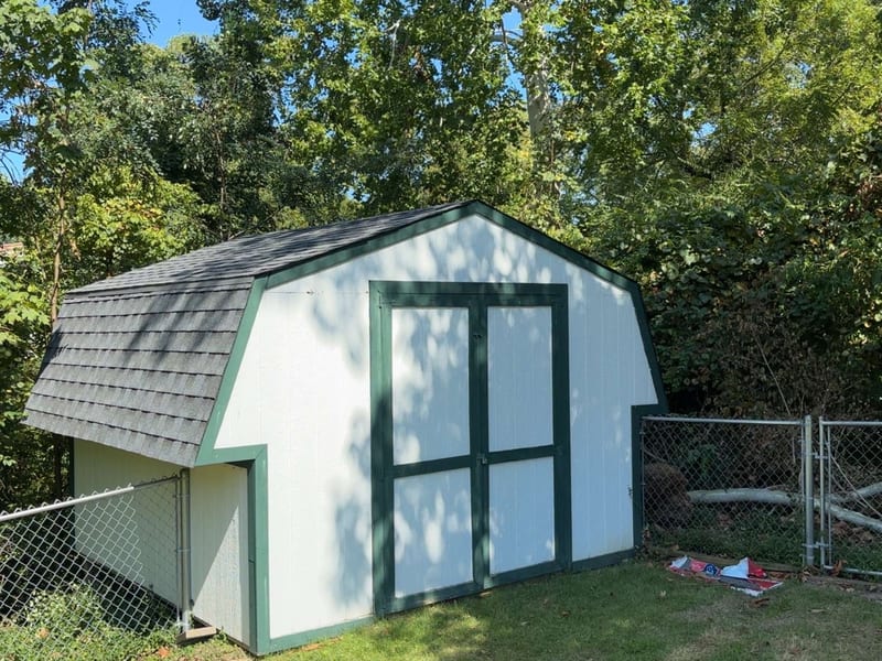 White gambrel shed with dark green trim and double doors under a new charcoal shingle roof, chain-link fence and woods behind