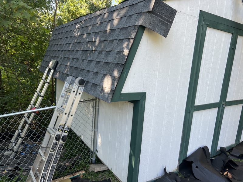 New charcoal shingles and ridge cap on the shed's gambrel slope, ladders leaning against the white siding, old black roofing piled below
