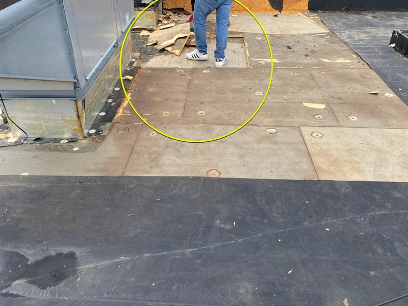 Exposed roof deck during an EPDM tear-off beside a rooftop exhaust unit, showing weathered wood-fiber board and rusted fastener plates as the crew re-fastens the deck before new insulation and membrane go down