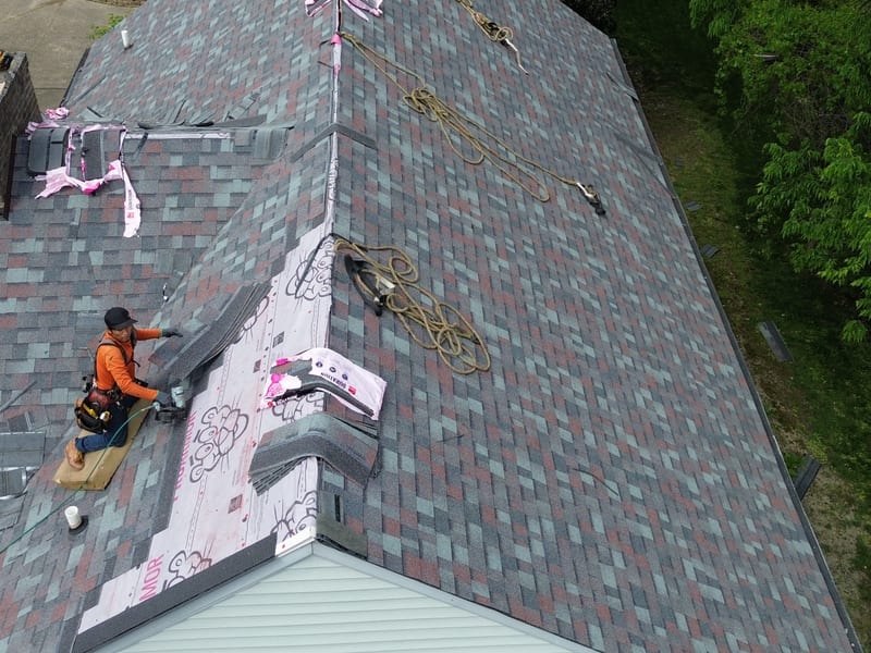 Roofer nailing hip cap where the new architectural shingle field meets the last strip of underlayment beside the chimney