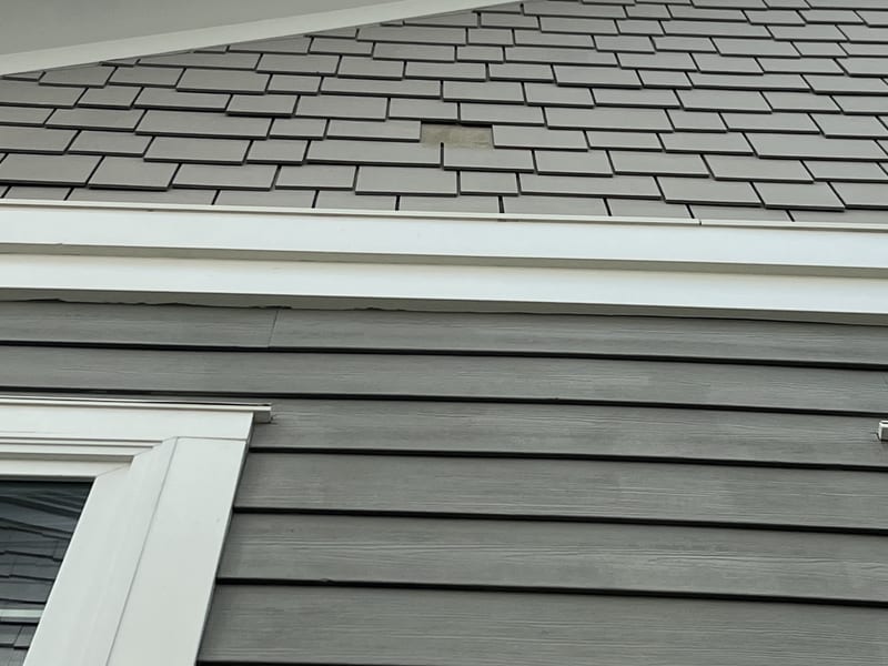 Front gable fiber-cement shake field with one shake missing, the bare patch and old adhesive residue visible mid-field