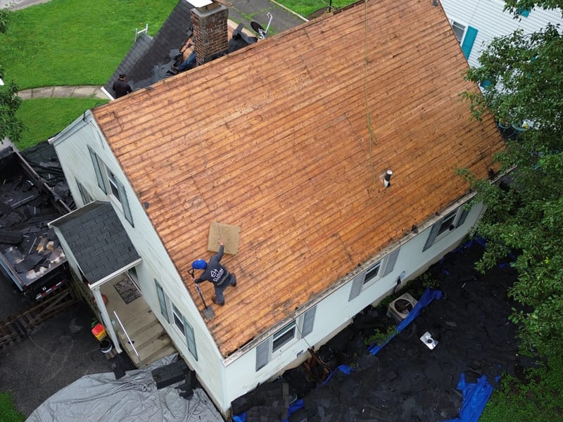 A roofer working the bare deck with a tear-off shovel, dump trailer loaded and tarps protecting the beds and AC unit