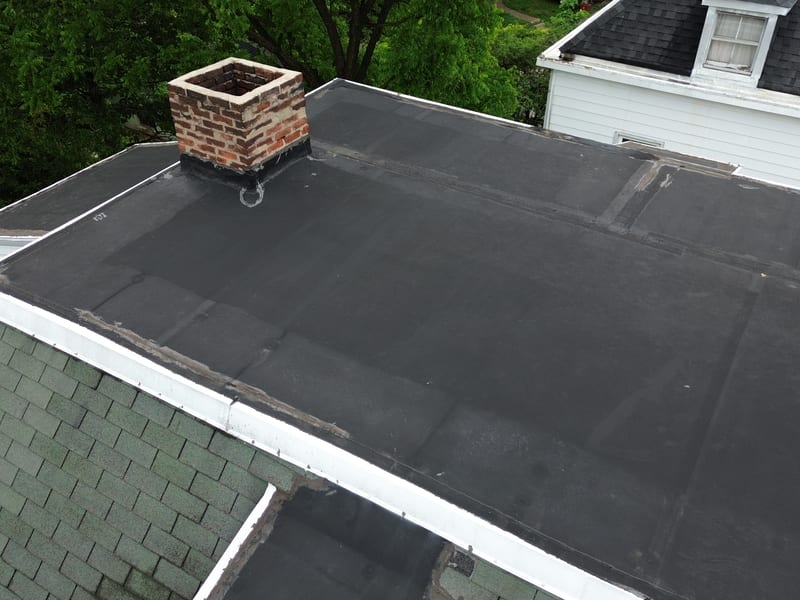 Angled view of the third-story flat roof with aging membrane, visible seams and patches, and the brick chimney rising off it
