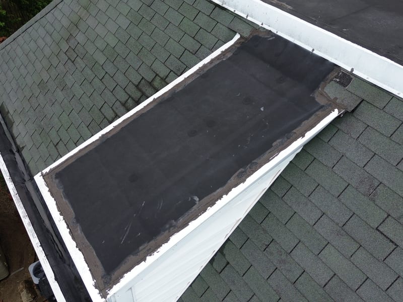 Top-down view of a bay cap where a rubber membrane panel sits in the surrounding aged shingle field, edges sealed with mastic