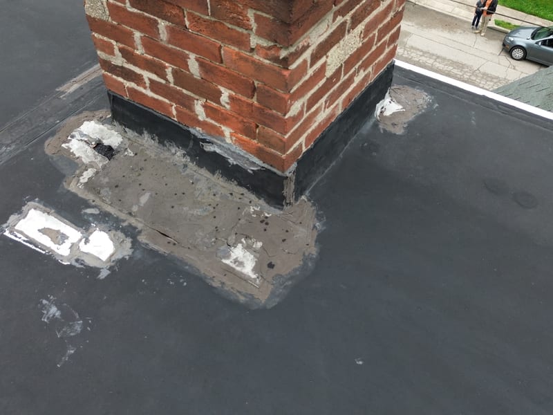 The brick chimney base on the flat roof buried under layers of troweled tar and sealant from prior repairs