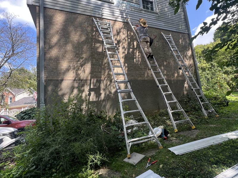 Three extension ladders set against the tall gable while a crew member installs new vinyl siding over fresh house wrap