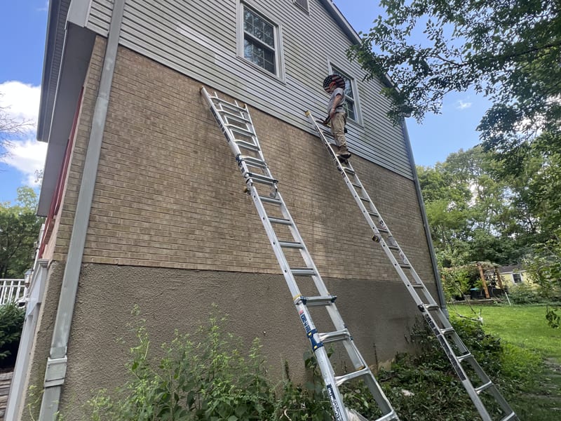 Crew member on an extension ladder fitting a replacement siding panel high on the gable