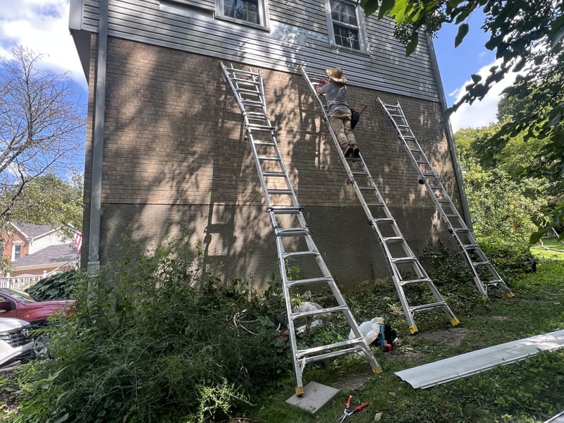 The damaged band stripped of siding panels — house wrap and brick exposed across the gable while the new panels wait below