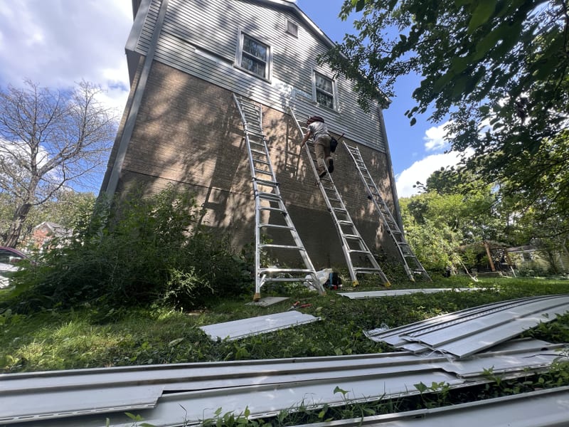 Three ladders on the stripped wall band with new siding panels staged and a crew member climbing