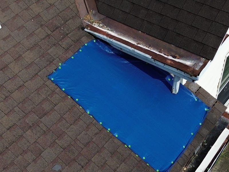 Aerial close-up of a blue emergency tarp secured to a shingle roof with tidy rows of green cap nails below a damaged eave